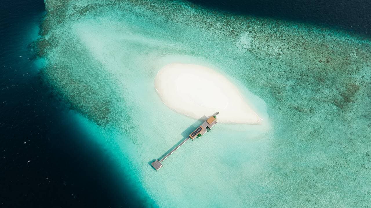 Aerial view of a Maldivian sandbank ringed by turquoise reef, with a jetty leading to a single overwater pavilion