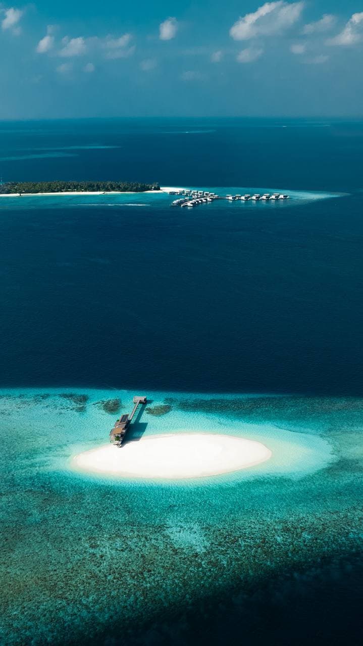 Aerial view of a sandbank in the Maldives, ringed by reef, with a resort island on the horizon