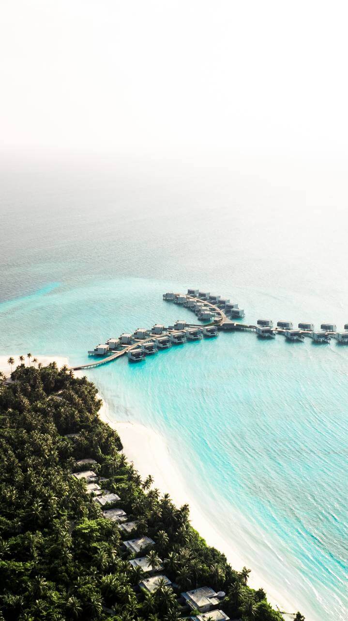 Aerial view of a Maldivian resort island, with a curving line of overwater villas reaching into a turquoise lagoon